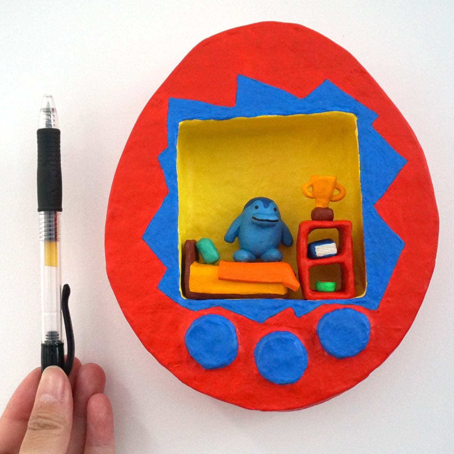 red tamagotchi inspired sculpture that has a blue character inside sitting on a bed with a small shelf next to it with a trophy on top next to a pen for scale reference